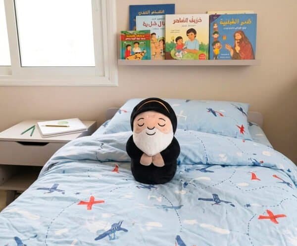 Saint Charbel plushie, a Maronite Catholic gift, sitting on a child's bed near a shelf of Lebanese books